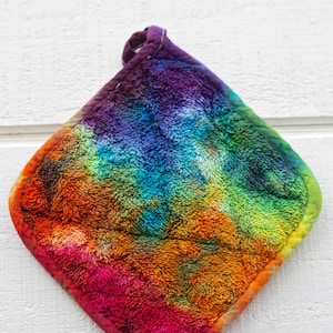 May include: A colorful, tie-dye pot holder with a loop for hanging. The pot holder features a vibrant spectrum of colors, including red, orange, yellow, green, blue, and purple. The edges are a darker shade of purple.