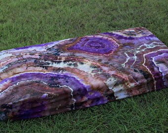 May include: A folded purple, brown, and white tie-dye fabric with a circular pattern. The fabric is folded and lying on a bed of green grass.