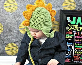 May include: A baby wearing a green and yellow crocheted dinosaur hat with a long tail. The hat has a pointed top and a wide brim. The baby is wearing a dark blue blazer and blue jeans.