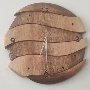 Peut inclure: Une horloge murale circulaire composée de segments de bois ondulés superposés, alternant entre des tons brun foncé et clair. Le cadran présente des chiffres gravés aux positions 12, 3, 6 et 9, des points creusés pour les autres heures et de fines aiguilles argentées.