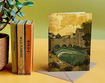 May include: A greeting card with a yellow and green illustration of a bridge over a river with buildings in the background. The card is on a yellow surface with three books stacked behind it.