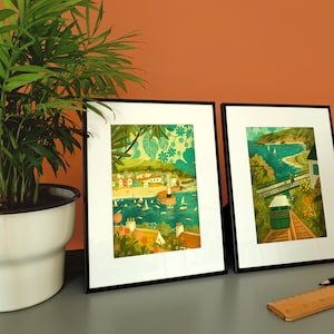 May include: Two framed illustrations of coastal scenes. The first shows a colorful seaside town with boats and a lighthouse. The second shows a green hillside with a cable car and a view of the ocean.