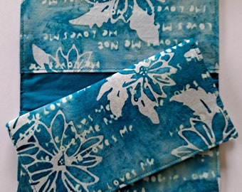 May include: A teal blue fabric wallet with a white floral pattern and the words "Loves Me" repeated on the front. The wallet is folded in half and the inside is lined with a solid teal blue fabric.