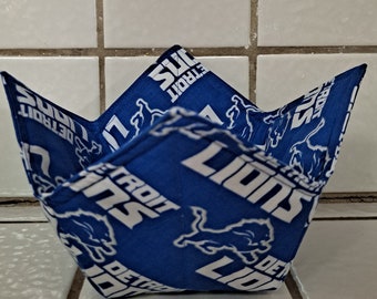 May include: A fabric bowl cosy featuring a repeating pattern of the Detroit Lions logo and text in white on a blue background. The square-shaped holder has four folded corners and stitched seams, designed to hold a bowl securely in place.
