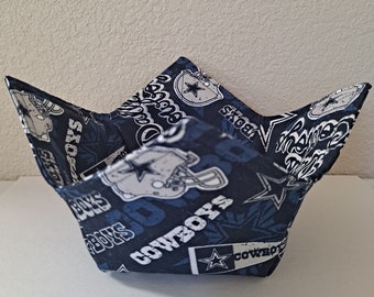 May include: A fabric bowl cosy featuring a dark blue background with a repeating pattern of Dallas Cowboys football helmets, stars, and the text Cowboys in white and grey. The square-shaped holder has four pointed corners.