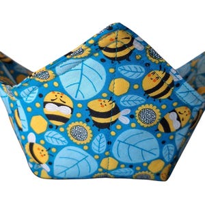 May include: A blue fabric bowl cozy with a repeating pattern of yellow bees, sunflowers, and light blue leaves. The cozy is designed to hold a bowl and has a folded, angular shape.