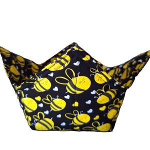 May include: A black and yellow bowl cozy with a bee and heart pattern. The cozy is made of fabric and has a unique shape with four pointed corners. The bees are yellow with black stripes and the hearts are white.