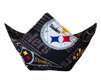 May include: A fabric bowl cosy featuring a black background with a repeating Pittsburgh Steelers logo pattern. The design includes the team name in yellow and white text, along with the iconic circular logo containing yellow, red, and blue stars.