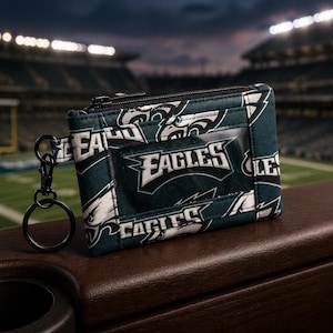 Może przedstawiać: Ciemnozielony portfel na identyfikator z tkaniny z powtarzającym się wzorem logo Philadelphia Eagles w kolorze białym, czarnym i srebrnym. Portfel posiada przezroczyste plastikowe okienko z przodu, czarny zamek błyskawiczny i czarny metalowy karabińczyk.