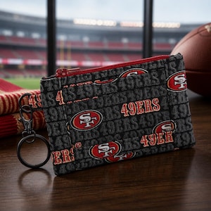 Puede incluir: Un estuche de tela negra con cremallera que presenta un patrón repetitivo del logotipo de los San Francisco 49ers y el texto 49ERS en rojo, dorado y blanco. El estuche incluye un mosquetón de metal negro y una anilla circular lateral.