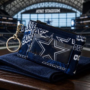 Puede incluir: Un monedero de tela azul marino con un patrón repetitivo del logotipo de estrella y texto de Dallas Cowboys en blanco. El estuche rectangular incluye una ventana de plástico transparente en el frente, una cremallera negra superior y un llavero de metal dorado.