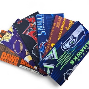 Op de afbeelding: Een verzameling stoffen portemonnees, elk met een ander teamlogo en -naam. De portemonnees tonen logo's voor de Seahawks, Washington, Tennessee en andere teams. De portemonnees zijn in verschillende kleuren, waaronder blauw, paars en oranje.