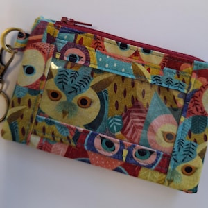 Puede incluir: Una pequeña bolsa con cremallera y una ventana de plástico transparente. La bolsa está hecha de tela con un estampado de búhos coloridos. La tela es una mezcla de rojo, amarillo, verde, azul y marrón. La bolsa tiene un llavero dorado.