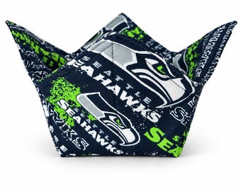 May include: A fabric bowl cosy featuring a dark navy blue background with a repeating Seattle Seahawks logo pattern in white, grey, and neon green. The bowl holder has four pointed corners and visible stitching along the edges and seams.