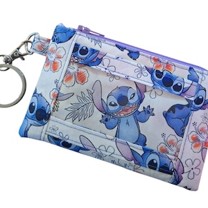 May include: A small, rectangular coin purse featuring a repeating pattern of a blue cartoon character with large ears and a floral design. The purse has a purple zipper, a silver key ring, and a silver clasp.
