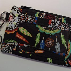 Könnte beinhalten: Schwarze Reißverschlusstasche mit einem bunten Traumfänger- und Federdruck. Die Tasche hat ein transparentes Plastikfenster und einen silbernen Schlüsselring.