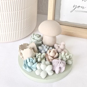 Könnte beinhalten: Ein hellgrüner Keramik-Ringhalter mit einem Pilz-Oberteil und pastellfarbenen Sukkulenten-Dekorationen. Der Ringhalter ist von einer weißen Oberfläche umgeben. Der Text "do what you love and love what you do" ist im Hintergrund sichtbar.