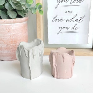 May include: Two concrete candle holders, one in a light gray color and the other in a light pink color. Both holders are shaped like candles with dripping wax detail. The holders are sitting on a white surface.
