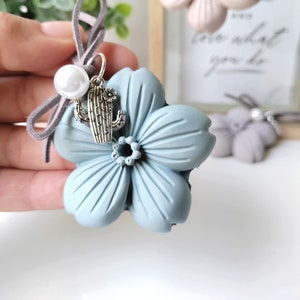 以下が含まれることがあります： 薄い青色のセラミックの花のチャームに、銀色のサボテンのチャームと真珠のビーズが付いています。チャームは茶色のスエードコードに取り付けられています。