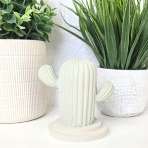 May include: A light green concrete cactus with a ribbed texture and two arms. The cactus is sitting on a white, tiered base.