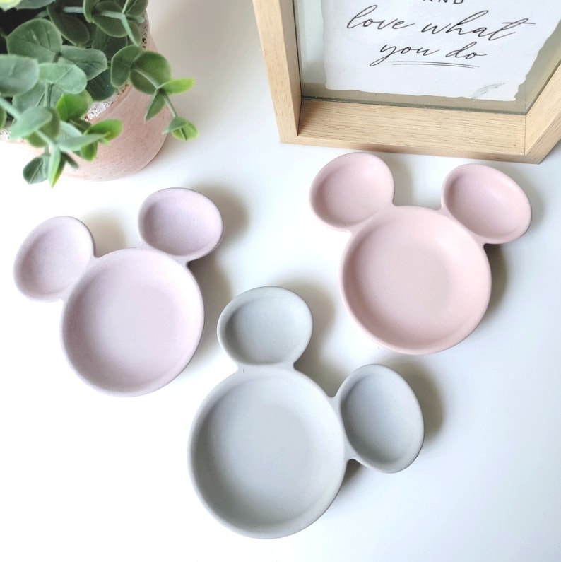 May include: Three ceramic dishes shaped like Mickey Mouse heads. The dishes are in pink and gray colors. The dishes are perfect for serving snacks or dips.