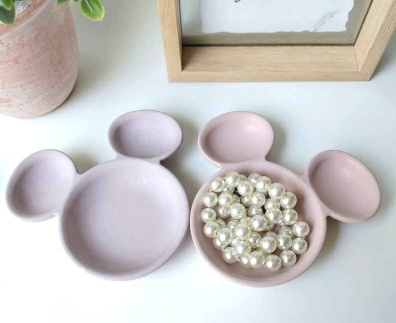 May include: Two pink ceramic dishes shaped like Mickey Mouse heads. One dish has a pearl necklace inside.