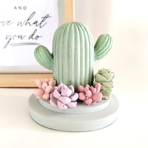 Puede incluir: Un cactus de hormigón verde con una base verde claro y tres suculentas rosas y moradas. El cactus tiene una textura acanalada y un solo brazo.