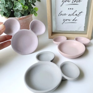 May include: Three ceramic dishes shaped like Mickey Mouse heads. The dishes are in pink, light pink, and gray.