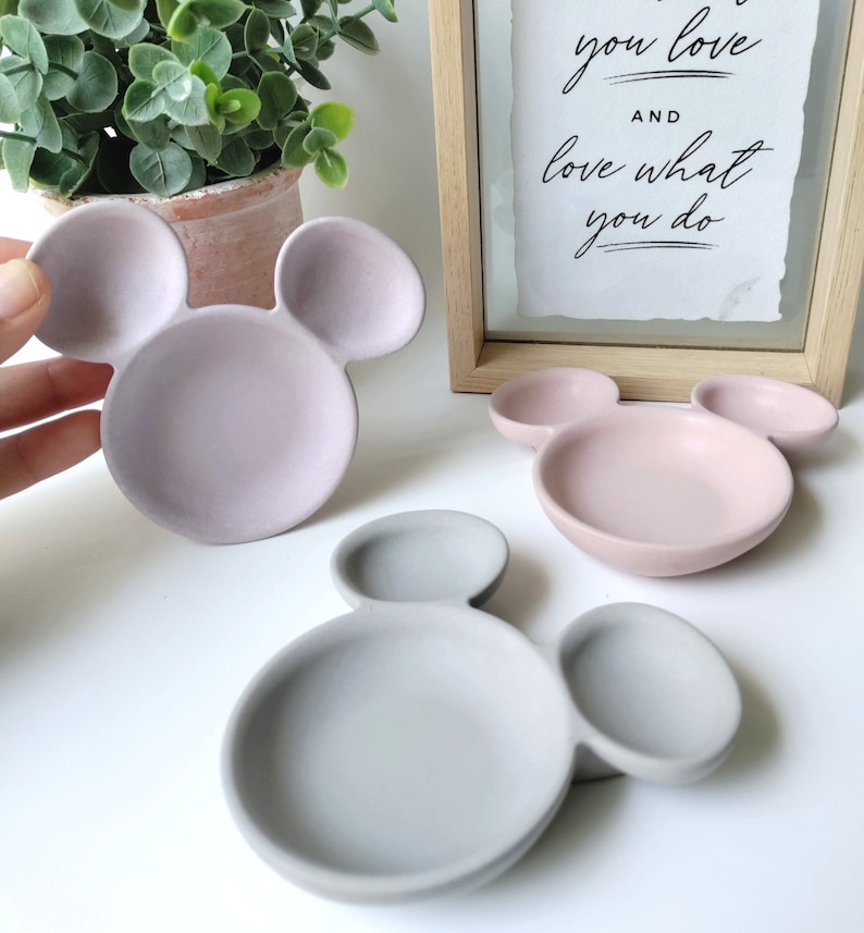 May include: Three ceramic dishes shaped like Mickey Mouse heads. The dishes are in pink, light pink, and gray.