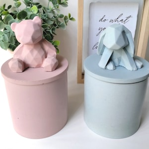 May include: Two ceramic containers with geometric animal figurines on top. The container on the left is pink with a bear figurine. The container on the right is light blue with a rabbit figurine. The text "do what you love" is visible on a framed sign behind the containers.
