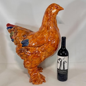 May include: A large, orange ceramic hen statue with detailed feather textures and a glossy finish. The hen is standing next to a dark wine bottle with a label featuring two geese.