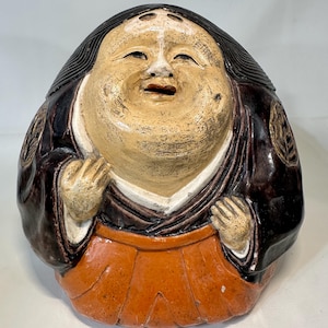 May include: A ceramic figure of a smiling, stout person with a light skin tone. The figure wears a dark brown jacket over a white shirt and an orange skirt. The figure's hair is black, and the hands are clasped near the chest.