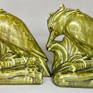 May include: A pair of green ceramic bird bookends. Each bookend features a bird perched on a branch with a curved beak and detailed feathers. The bookends are a glossy, olive green color.