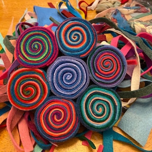 Puede incluir: Una colección de rollos de fieltro coloridos, cada uno con un patrón en espiral. Los rollos están dispuestos en un círculo, con los extremos de las tiras de fieltro que se extienden desde el centro. El fieltro es de varios colores, incluyendo rojo, azul, verde, amarillo y morado.