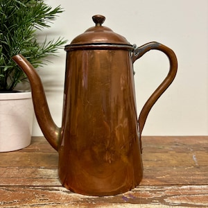 May include: A vintage copper coffee pot with a lid and curved handle. The pot has a long spout and is set on a wooden surface. The copper has a warm, reddish-brown hue, suggesting age and use. A small green plant is visible in the background.
