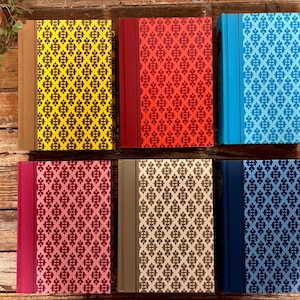 May include: A collection of six hardcover books with patterned covers in various colors, including yellow, red, and blue. The covers feature a repeating geometric design. The books are arranged on a wooden surface.