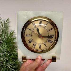 May include: A square, vintage clock with a gold-tone face and Roman numerals. The clock is set in a white marble-like frame with gold-tone supports. The hands indicate the time is approximately 10:15.