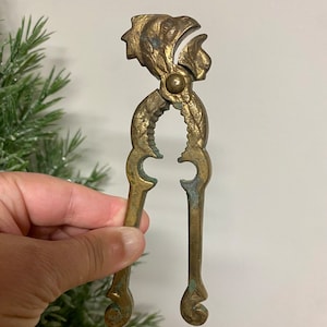 May include: A brass nutcracker shaped like a rooster with a detailed design. The nutcracker has a hinged jaw and a spring mechanism.