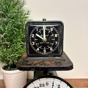 May include: A vintage black metal Gilbert interval timer with a square face and white numerals sits atop a black metal scale. The scale features a round white dial with black markings and text reading 24 Lbs. Oz. and a black metal pointer.