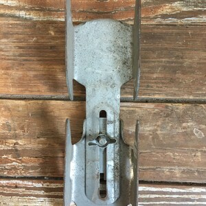 May include: A pair of vintage metal ice skate holders. The holders are silver in color and have a simple design. They are attached to a wooden surface.