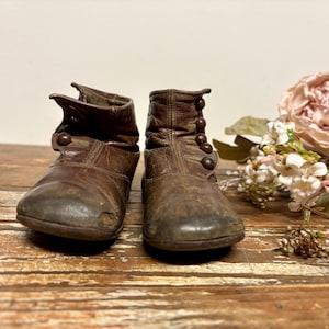 May include: A pair of weathered brown leather ankle boots featuring a rounded toe, a side-button closure with four dark circular buttons, and visible signs of wear on the front toes. The boots have a structured shaft and a low profile design.