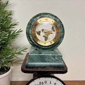 May include: A green marble clock with a gold-colored face featuring a world map design. The clock sits on a matching green marble base and a dark brown surface. The clock face has black numbers and hands.