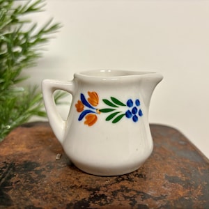 May include: A small, white ceramic creamer with a handle and spout. The creamer is decorated with orange, blue, and green floral designs. It sits on a rustic, brown surface.