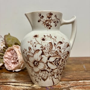 May include: Cream-colored ceramic pitcher with a brown floral pattern. The pitcher has a handle and a spout. The floral design includes roses and other flowers. The pitcher is on a wooden surface.