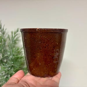 May include: A small, cylindrical ceramic pot with a dark brown, speckled, glossy glaze. The vessel features a slightly flared rim and a smooth, uniform texture throughout its exterior surface, showcasing a deep, earthy tone with subtle light flecks.