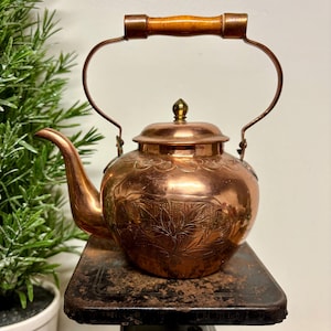 May include: A copper tea kettle with a wooden handle and floral engravings. The kettle has a curved spout and a lid with a small decorative knob. The kettle is sitting on a vintage scale.