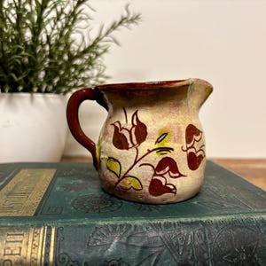 May include: A small ceramic creamer pitcher with a cream-colored base, featuring hand-painted dark brown floral motifs and green leaf accents. The pitcher has a curved handle, a small pouring spout, and a dark brown rim around the top edge.
