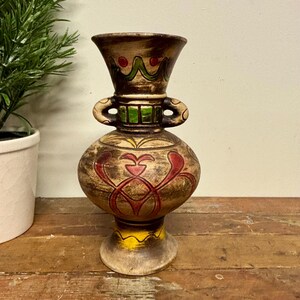 May include: A vintage, handcrafted ceramic vase with a bulbous body and flared top. The vase features brown, red, green, and yellow painted details. It has two loop handles and a pedestal base. The vase is approximately 10 inches tall.