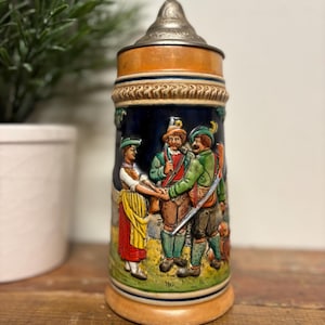 May include: A ceramic beer stein with a pewter lid. The stein features a colorful scene of three people in traditional German clothing. The scene is set against a blue background with a white border.