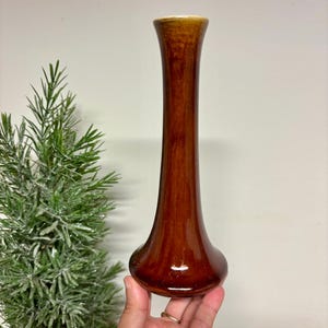 May include: A tall, slender ceramic bud vase with a glossy, deep brown glaze. The vase features a flared base that tapers into a narrow neck, widening slightly at the top rim. The surface has a smooth, reflective finish with subtle color variations.